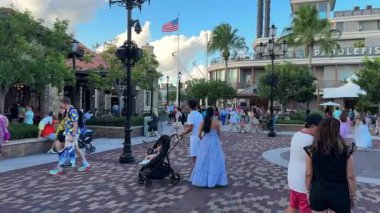 Orlando, Florida, ABD - 08.31.2024: İnsanlar dükkanlar, restoranlar ve ağaçlarla çevrili işlek tuğla bir meydanda yürüyor. Sahnenin üzerinde dalgalanan bir Amerikan bayrağı Parlak mavi bir yaz gökyüzünün altında.
