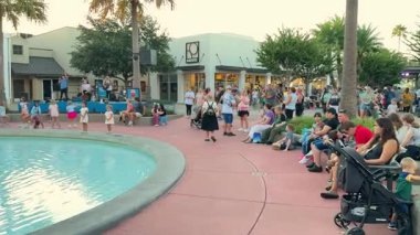 Orlando, Florida, ABD - 08.31.2024: Bir grup Orlando, Florida 'daki Disney Springs sahnesinde konser verirken aileler kavisli bir çeşmenin etrafında oturur ve ayakta dururlar. Kalabalık açık hava gösterisinin keyfini çıkarırken çocuklar su kenarında dans edip oynuyorlar..