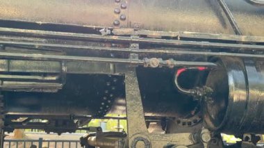 Boruların, çubukların ve metal bileşenlerin yakın görüntüsü eski siyah buharlı bir lokomotifin yanından geçiyor. Yıpranmış boya ve perçinler tarihi trenin yaşını ve endüstriyel karakterini gösteriyor..
