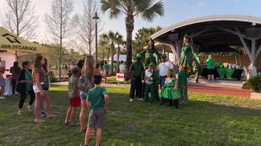 Orlando, Florida, ABD - 03.16.2024: Kostümlü Aziz Patrick Günü sanatçıları ve leprikonları güneşli bir Florida parkında aileleri ve çocukları ile poz verdiler. İnsanlar sahnenin yanında gülümsüyor, palmiye ağaçları ve satıcı çadırları, neşeli bir bayram festivali sahnesi yaratıyor..