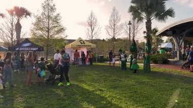 Orlando, Florida, ABD - 03.16.2024: Aileler ve sanatçılar, satıcı çadırları ve palmiye ağaçlarıyla bir Aziz Patrick Günü festivalinde çimlerin üzerinde toplandılar. Yeşil elbiseli insanlar, güneşli havanın, eğlencenin ve deniz kenarındaki topluluk aktivitelerinin keyfini çıkarıyorlar..