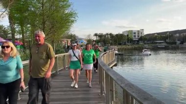 Orlando, Florida, ABD - 03.16.2024: Toplum etkinliği sırasında yeşil tişört giyen insanlar göl kıyısında yürüyorlar. Grup sıcak akşam ışığında rahatça rıhtımda geziniyor..