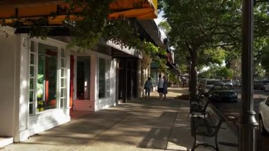 Winter Park, Florida, ABD - 03.30.2024: Gün ışığı butik mağazalarının vitrinlerine ve ağaçlı bir alışveriş caddesindeki kaldırım banklarına düşer. İnsanlar küçük bir kasaba merkezinde park halindeki arabaların, tentelerin ve vitrinlerin önünden geçiyor..