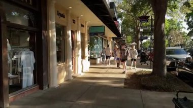 Winter Park, Florida, ABD - 03.30. 2024: İnsanlar butik mağazalarının ve giysi vitrinlerinin önünden geçen güneşli bir kaldırım boyunca yürüyorlar. Park edilmiş arabalar, gölgelik ağaçlar ve vitrinler küçük bir alışveriş caddesi oluşturur..
