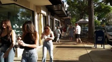 Winter Park, Florida, ABD - 03.30. 2024: Günlük kıyafetler içindeki genç kadınlar dondurma yerken işlek bir alışveriş caddesi boyunca yürürler. Mağaza vitrinleri, park halindeki arabalar ve ağaçlar sıcak güneş ışığında canlı bir kaldırım sahnesi oluşturuyor..