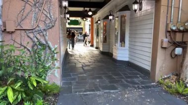 Winter Park, Florida, ABD - 03.30.2024: Sallanan fenerlerle vitrinler arasındaki dar yaya geçidi. İnsanlar taştan koridorda güneşli sokağa doğru yürüyorlar..