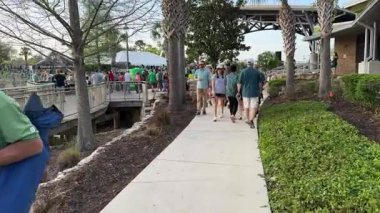 Orlando, Florida, ABD - 03.16.2024: Sıradan yeşil elbiseli insanlar kalabalık bir açık hava festivali sırasında kaldırım boyunca yürürler. Palmiye ağaçları, çimenler, tahta kaldırımlar ve çadırların yakınındaki kalabalık sıcak bir şehirde sosyal bir tatil etkinliği gösteriyor..