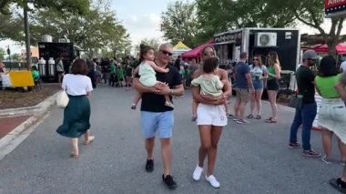 Orlando, Florida, ABD - 03.16.2024: Ebeveynler yiyecek kamyonlarıyla dolu kalabalık bir festival caddesinde yürürken küçük çocukları taşıyor. Yaz kıyafetleri ve yeşil gömlekli insanlar açık hava tatilinin tadını çıkarıyorlar..