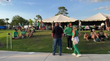 Orlando, Florida, ABD - 03.16.2024: Yeşil gömlekli insanlar büyük beyaz bir festival çadırının yanındaki çimenli bir tepede oturuyorlar. Akşam ışığı ve rahat gruplar açık havada dostça bir tatil ortamı yaratıyor..