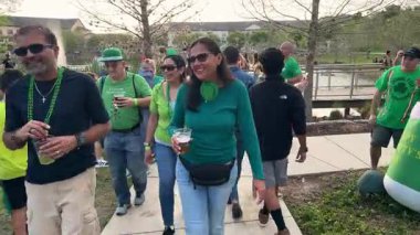 Orlando, Florida, ABD - 03.16.2024: St. Patricks Günü kutlamaları sırasında yeşil gömlekli ve aksesuarlı gülümseyen yetişkinler kalabalık bir parkta yürürler. İnsanlar canlı festivalde göl kenarında içki içip gülüyorlar..