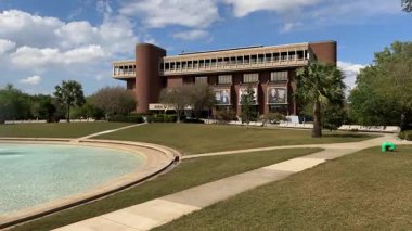 Orlando, Florida, ABD - 03.18.2024: Tuğla üniversite kütüphanesinin önündeki Üniversite Çeşmesi. Yeşil bir çimenlik, palmiye ağaçları ve bulutlu mavi gökyüzü parlak bir akademik kampüs sahnesi yaratır..