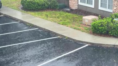 Boş bir otoparka ve binanın yanındaki ıslak kaldırıma sağanak yağmur yağıyor. Çimenler ve çalılar fırtınada sırılsıklam olurken, su karanlık asfaltta süzülür..