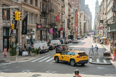 New York City, New York, ABD - 08.01.2024: Manhattan 'da kalabalık bir caddenin köşesinde insanlar, dükkanlar ve tarihi binalarla çevrili sarı bir taksi dönüyor. Moda mağazaları ve etkin yaya trafiği New York gününü yansıtıyor.
