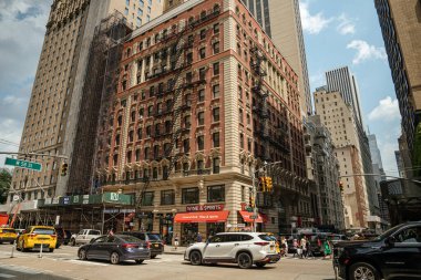 New York City, New York, ABD - 08.01.2024: Dekoratif mimarisi ve metal yangın merdivenleri olan klasik bir kırmızı tuğla bina hareketli bir New York sokak köşesinde duruyor. Sarı taksiler ve yayalar şehir merkezinde güneşli bir gökyüzünün altında kavşağı geçiyorlar.