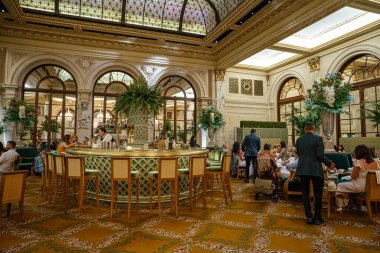 New York City, New York, ABD - 08.01.2024: Sofistike bir otel salonu, içki ve sohbetten keyif alan misafirlerle çevrili merkezi bir bara sahiptir. Süslü iç mekan vitrinleri kemerli pencereler, çiçek aranjmanları ve zengin dekore edilmiş bir tavan..