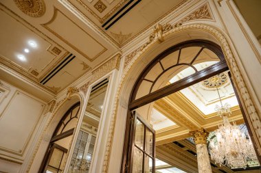 Zarif kemerli bir kapı, avizeler, altın sütunlar ve klasik tavan detaylarıyla süslenmiş büyük bir odaya açılıyor. Zengin dekore edilmiş mimari tarihi lüks ve karmaşıklığı yansıtıyor..