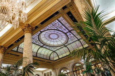 Lüks bir otel lobisinin üzerinde mermer sütunlar ve bereketli iç mekan bitkileriyle güzel bir vitray kubbe. Klasik mimari ve karmaşık detaylar zarafet ve ihtişam ortamı yaratır..