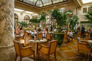 New York City, New York, ABD - 08.01.2024: Konuklar mermer sütunlar, tropikal bitkiler ve süslü dekorlarla çevrili lüks bir otel salonunda çay ve sohbetin keyfini çıkarıyorlar. Mekanda vitray tavan, çiçek aranjmanları ve klasik