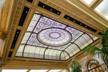 Lüks bir otel lobisinde çiçekli ve geometrik desenli süslü cam bir kubbenin ayrıntılı görüntüsü. Zarif pervazlar, mermer sütunlar ve bereketli yeşillik klasik ortamı tamamlıyor.