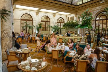 New York City, New York, ABD - 08.01.2024: İnsanlar mermer sütunları, yemyeşil bitkileri ve boyalı cam tavanı olan lüks bir otel salonunda zarif bir öğleden sonra çayının keyfini çıkarıyorlar. Odada klasik mobilyalar, çiçek aranjmanları ve sıcak konukseverlik bulunuyor..