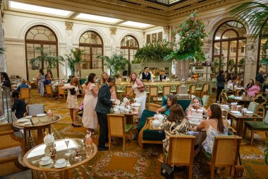 New York City, New York, ABD - 08.01.2024: Konuklar şık bir otel salonunda toplanarak mermer sütunlar, çiçek dekoru ve yemyeşil alanlar arasında çay servisi yapıyorlar. Kaliteli iç mekan klasik mobilyalar, vitray ayrıntıları ve rafine edilmiş