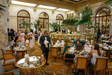 New York City, New York, ABD - 08.01.2024: Resmi giyinmiş bir garson zarif bir otel salonunda öğleden sonra çayının keyfini çıkaran misafirlere servis yapıyor. Etrafında mermer sütunlar, çiçek aranjmanları ve klasik dekorlarla çevrili olan dekor, zamansız karmaşıklık yayar..