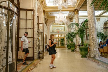 New York City, New York, ABD - 08.01.2024: İnsanlar avizeler, mermer kolonlar ve tropikal bitkilerle süslenmiş büyük bir otel lobisinde yürüyor. Klasik dekor ve dekoratif mimari iç mekanın lüksünü vurgular..