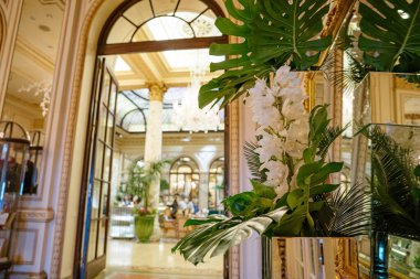 Tropikal yapraklı beyaz bir çiçek aranjmanı lüks bir otel odasının girişini süslüyor. Aynadaki yansımalar mermer sütunlar, avizeler ve içinde vitraylı bir tavan olduğunu gösteriyor..
