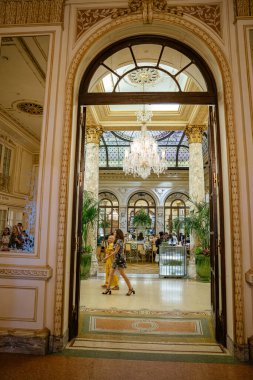 New York City, New York, ABD - 08.01.2024: Süslü bir koridor yüksek tavanları, mermer sütunları, avizeleri ve boyalı cam tavan penceresi olan Grand Plaza Otel yemek alanına çıkıyor. Gösterişli konuklar, kapalı alandaki yemyeşil bitkilerin ve klasik mimarinin yanından geçiyor.