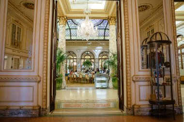 New York City, New York, ABD - 08.01.2024: Süslü bir Plaza otel lobisi mermer kolonlar, avizeler ve yemyeşil bitkilerle lüks bir yemek odasına çıkıyor. Konuklar, New York City 'deki cam tavan altında rafine bir atmosferin keyfini çıkarıyorlar.