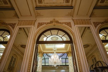 Süslü kemerli bir kapı büyük bir avizeye açılıyor ve mermer sütunlarla çerçevelenmiş cam bir tavan. Ayrıntılı pervazlar, altın aksanlar ve mimari simetri tarihi bir New York otelinde klasik lüksü yansıtır..