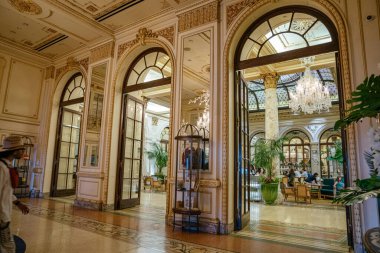 New York City, New York, ABD - 08.01.2024: Tarihi bir New York otel lobisi uzun kemerli kapılar, mermer sütunlar ve rafine bir yemek alanına açılan kristal avize içerir. Konuklar, yemyeşil bitkilerle çevrili lüks bir ortamda rahatlar.