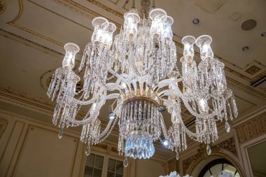 Süslü bir kristal avizenin detaylı görüntüsü tarihi lüks bir otelin tavanından ışıldıyor. Cam damlacıklar, karmaşık şekiller, ve klasik iç dekorda şık ışıklandırma zenginliği ve zanaatkarlığı..
