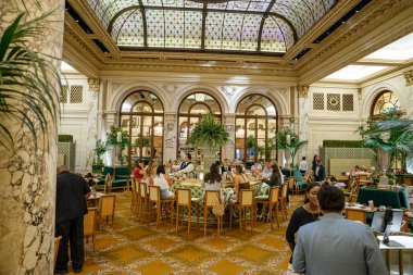 New York City, New York, ABD - 08.01.2024: Tarihi bir New York otel salonunun vitray tavanının altındaki merkez barın etrafında şık bir kalabalık toplanıyor. Mermer sütunlar, kadife koltuklar ve yemyeşil çiçeklerle çevrili misafirler,