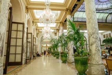 New York City, New York, ABD - 08.01.2024: Bir grup kadın avizeler, mermer sütunlar ve uzun yeşil bitkilerle süslenmiş bir otel lobisinde yürüyor. Alan kemerli cam kapılar, altın tavan aksanları ve zarif