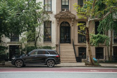 Dekoratif detaylarla dolu lüks bir apartman girişi park edilmiş bir cipin arkasında duruyor. Ağaçlar ve kaldırımlar rafine edilmiş bir şehir manzarası..