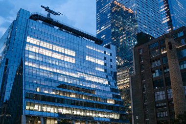 Manhattan 'da cam bir ofis binası gece aydınlanıyor ve üzerinde inşaat vinci var. Sahne, New York 'taki kentsel gelişim ve kurumsal büyümeyi yansıtıyor.