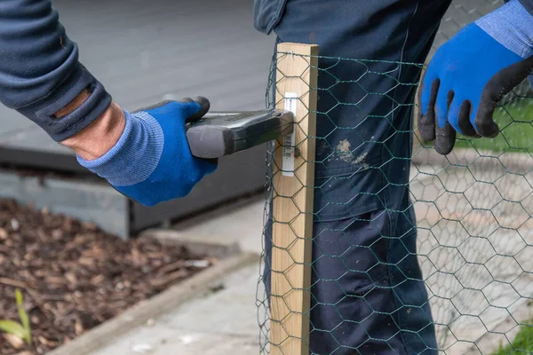 Zımba erkek çivileri kullanarak ahşap kazığa tel örgü örüyorlar. DIY projesi arka bahçede.