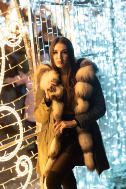 Kürk manto, mini etek ve turuncu kazak giyen koyu renk saçlı güzel bir kız. Gülümsüyor, arka planda ışıklarla poz veriyor.