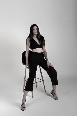 Siyah bluzlu ve pantolonlu hoş bir esmer sandalyede oturuyor. Beyaz arkaplan