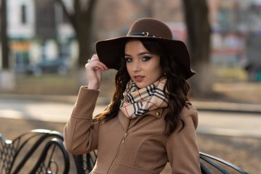 Kahverengi paltolu ve şapkalı güzel genç kız şehir parkında bir bankta oturuyor.