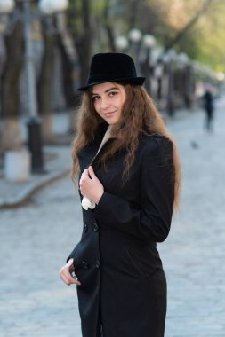 Siyah paltolu ve siyah şapkalı güzel bir kadın şehir caddesinde poz veriyor. Kız yürüyor ve bahar gününün tadını çıkarıyor.