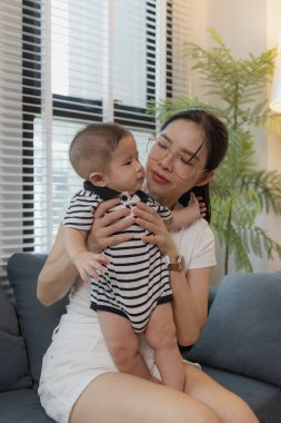 Genç bir anne, bebeğini sevgiyle kucaklar ve bir kanepede emzirir. Erken çocukluk ve aile hayatının anne bakımını, bağlarını ve besleyici anlarını sembolize eder..