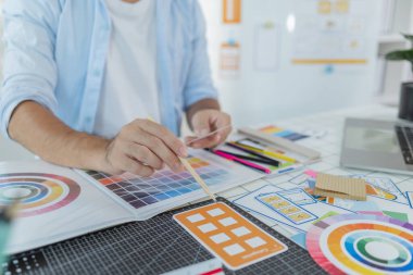 Bir tasarımcı masadaki yaratıcı bir UX UI projesi üzerinde çalışıyor, renk paletleri ve düzen şablonlarını odaklanma ve hassasiyetle düzenliyor, modern tasarım ve yeniliği temsil ediyor.