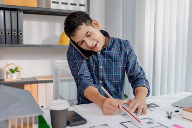 Genç bir mimar masasında birden fazla iş yapıyor, planlar çiziyor, telefon görüşmeleri yapıyor ve notları gözden geçiriyor. Çalışma alanı odaklanmayı, üretkenliği ve modern tasarım çalışmalarını gösteriyor..
