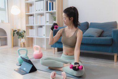 Genç bir kadın evde egzersiz yapıyor, yoga yapıyor ve halterleri ve dambılları ile egzersiz yapıyor. Egzersiz rutinini odaklanma ve kararlılıkla takip ediyor..