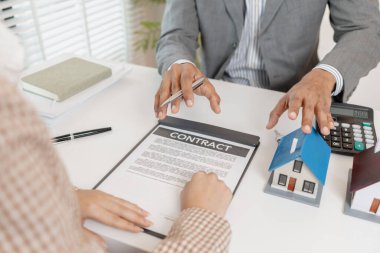 Bir emlakçı ve müşteri emlak satın alma seçeneklerini tartışıyor, sözleşmeyi gözden geçiriyor, kredi şartlarını hesaplıyor ve ev modellerini konut yatırımı kararının bir parçası olarak değerlendiriyor..