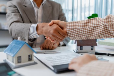 İş kıyafeti giyen iki kişi emlak anlaşmasını sonuçlandırıyor, ev modelleri, anahtarlar ve bir sözleşme ile el sıkışıyor, anlaşma, güven ve başarılı pazarlığı sembolize ediyor.