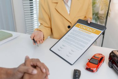 Sarı ceketli bir kadın satıcının, erkek bir müşteriye araba modellerini göstermesi ve anahtarları teslim etmesi ile ilgili detayları açıkladığı bir ofiste bir araba satışı pazarlığı gerçekleşiyor..