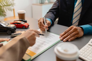 Bir işadamı ve müşteri, küçük model arabalar, bir kahve fincanı ve etraflarında araç satın alma pazarlığını sembolize eden belgeler içeren bir araba satış sözleşmesini inceliyor ve imzalıyor..